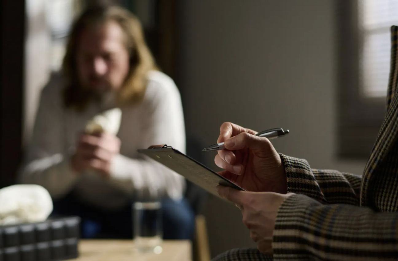 A person sitting in a counseling office while a hand holds a pen in the foreground.